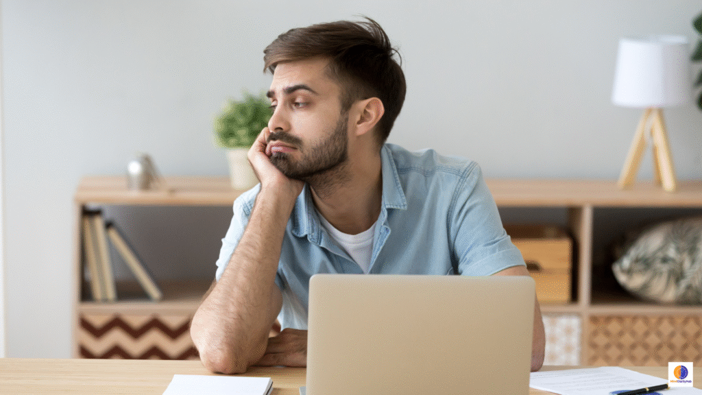 Adult sitting at a desk at dusk, staring past a glowing laptop with a distant expression, representing the quiet crisis of feeling unfocused and lost in modern life