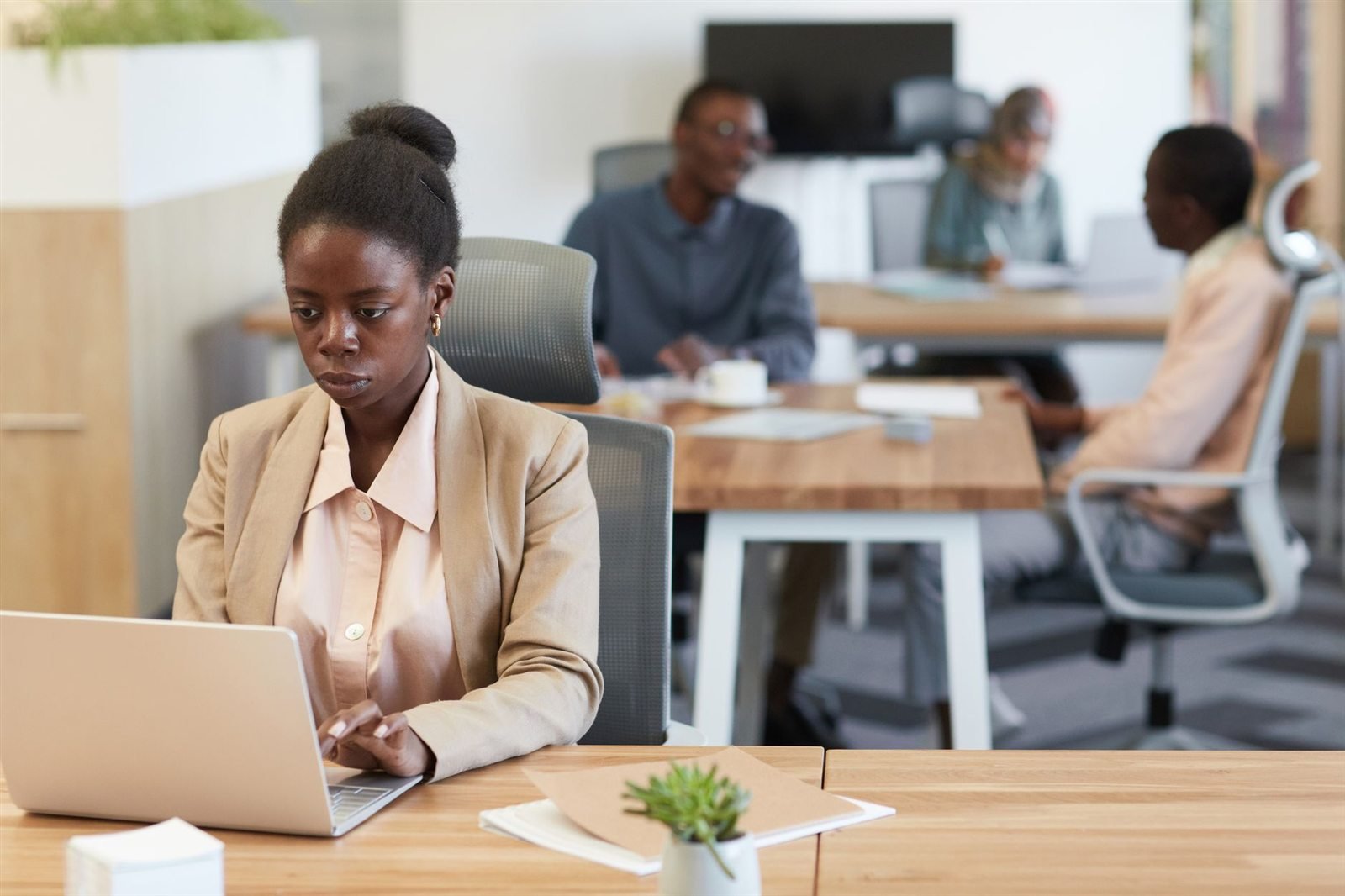A woman working at a laptop in the foreground while colleagues hold a meeting behind her